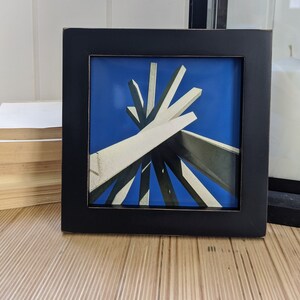 May include: A black framed picture with a blue background and a white and gray abstract sculpture in the center.