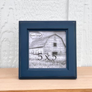 May include: A dark blue wood picture frame with a 5x5 inch opening. The frame is handmade in the USA and features a rustic design. The frame is sitting on a light wood surface.