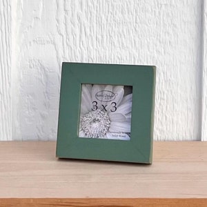 May include: A small, square, green picture frame with a white flower photo inside. The frame is made of solid wood and measures 3x3 inches.