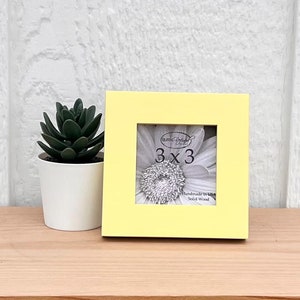 Puede incluir: Un marco de fotos amarillo de madera maciza de 7,6 x 7,6 cm. El marco contiene una flor blanca con un centro plateado.