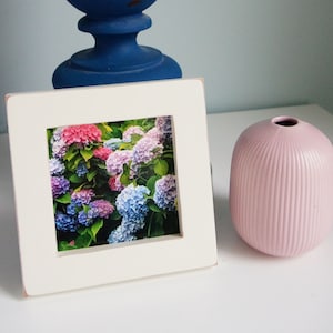 May include: White picture frame with a photo of a cluster of pink, blue, and purple hydrangeas. The frame is sitting on a white surface next to a pink ceramic vase.