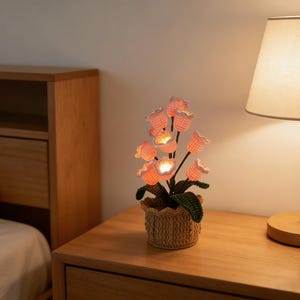 Puede incluir: Una lámpara de tulipanes de ganchillo con flores rosas iluminadas en una cesta tejida. La lámpara está sobre una mesita de noche de madera junto a una lámpara con una pantalla beige. La lámpara es una pieza decorativa.