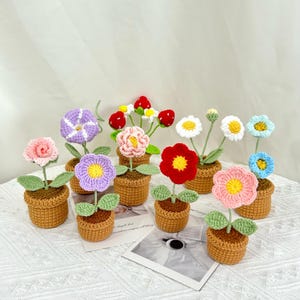 May include: A collection of colorful crochet flowers in small brown pots. The flowers vary in color, including pink, purple, red, and blue, with green leaves. The pots are arranged on a white surface, creating a charming, handcrafted display.