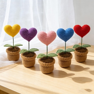 May include: Five crochet heart-shaped flowers in small brown pots. The hearts are yellow, purple, pink, blue, and red. Each flower has a green stem and leaves. The pots are on a light brown wooden surface.
