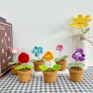 Pode incluir: Uma coleção de flores de crochê feitas à mão em pequenos vasos marrons. As flores são de várias cores, incluindo rosa, vermelho, azul, laranja e roxo, com folhas verdes. Uma flor amarela está em um vaso branco. O arranjo está em uma superfície xadrez.