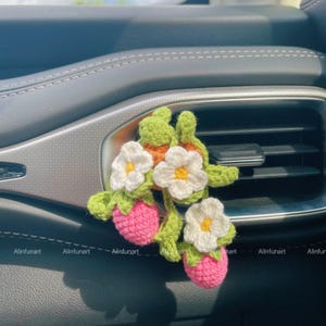 May include: A handmade crochet car air vent clip featuring a strawberry design. The clip has pink strawberries, white flowers with yellow centers, and green leaves. The clip is attached to a car's air vent.