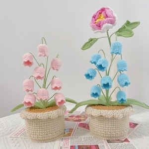 May include: Two crochet flower arrangements in beige pots. One features pink bell-shaped flowers, the other has blue bell-shaped flowers and a large pink and white flower. The pots have a scalloped edge.