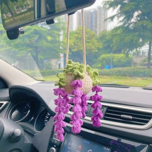 May include: A handmade crochet car charm featuring a hanging plant design. The charm has a beige pot with green leaves and cascading purple flowers. It hangs from a beige string, adding a touch of nature to the car's interior.