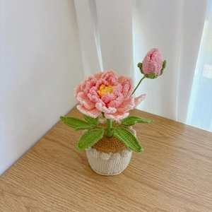 May include: A handcrafted crochet flower arrangement featuring a large, multi-layered pink peony with a yellow center and a single closed pink bud. The plant has green leaves and is set in a small, textured, cream-colored crocheted pot.
