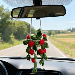Könnte beinhalten: Ein handgefertigter Autoanhänger aus Häkelarbeit mit roten Beeren und grünen Blättern. Der Anhänger ist an einer cremefarbenen Kordel aufgehängt und hängt am Rückspiegel eines Autos. Der Anhänger ist ein dekorativer Artikel.