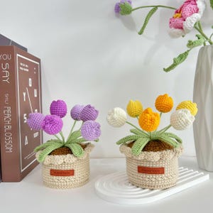 May include: Two handmade crochet tulip flower arrangements in beige pots. One arrangement features purple tulips, the other yellow and white. Each pot has a scalloped edge and a "Handmade" tag. The background includes a book and a white vase.