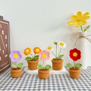 May include: A collection of handmade crochet flowers in small brown pots. The flowers are various colors, including purple, orange, pink, white, and red, with yellow centers and green leaves. A single yellow flower is in a white vase.