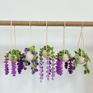 Puede incluir: Una colección de cinco macetas colgantes hechas a mano con ganchillo. Cada una presenta una maceta color crema llena de hojas verdes y flores de glicinas moradas. Las macetas están suspendidas de una barra marrón con cuerdas color crema.