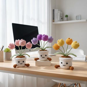 May include: Three crochet tulip flower arrangements in white pots with smiling faces and brown feet. The tulips are in shades of pink, purple, yellow, and orange. The arrangements are on a wooden desk with a computer in the background.