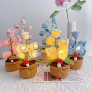 May include: Crocheted flower arrangements in pink, yellow, and blue, each with a small mushroom accent. The flowers are in brown woven pots. The flowers have small lights inside. The arrangement is on a white surface.