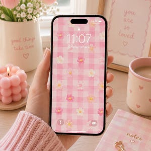 Puede incluir: Un teléfono inteligente que muestra un fondo de pantalla digital con un patrón de cuadros vichy rosa y blanco decorado con pequeñas flores dispersas de estilo acuarela. La pantalla muestra la hora 11:07 y la fecha domingo 18 de mayo.