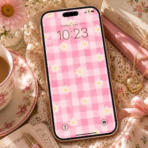 May include: A smartphone screen shows a pink gingham pattern with white daisy accents. The screen displays "Tuesday, May 14" and the time "10:23". The phone is on a lace surface, near a teacup, books, and a floral pendant.