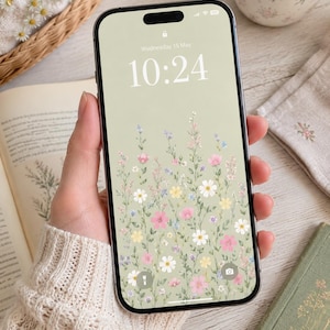 May include: A smartphone displaying a lock screen with a sage green background featuring a pattern of small pink, white, and yellow wildflowers. The screen shows the date Wednesday 15 May and the time 10:24 in a white, thin, sans-serif font.
