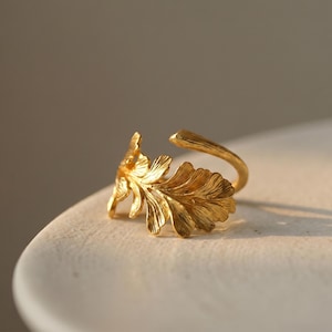 Può includere: Un anello color oro caratterizzato da un design dettagliato e testurizzato a forma di foglia di quercia che avvolge il dito. La superficie metallica presenta una finitura lucida e riflettente con solchi profondi che definiscono le venature.
