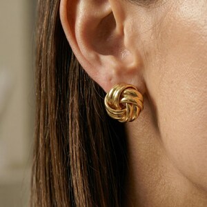 May include: Close-up of a gold-colored knot stud earring. The earring is round with a woven design. The earring is on a person's ear, with brown hair visible.