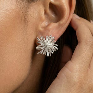 May include: A close-up of a silver stud earring in the shape of a spiky flower. The earring is worn on the ear of a person. The earring has a textured surface and a detailed design, reflecting light.