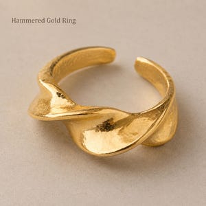 Op de afbeelding: Een gouden ring met een gehamerde textuur en een open, gedraaid ontwerp. Het oppervlak van de ring heeft een reflecterende, metalen afwerking met zichtbare inkepingen over de band. De tekst "Hammered Gold Ring" is zichtbaar in de linkerbovenhoek.