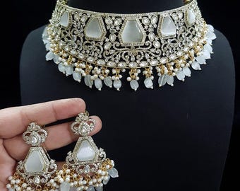 May include: An ornate silver-toned necklace and matching earrings set. The necklace features large, light-colored geometric stones, surrounded by intricate detailing and small, dangling beads. The earrings mirror the necklace design, with similar stones and bead accents.