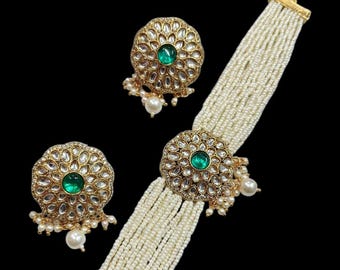 May include: A gold-toned jewelry set featuring a choker and matching earrings. The choker is made of multiple strands of small, white beads, with gold-toned circular accents. The earrings and choker accents have a green gemstone center and are surrounded by small, clear stones.
