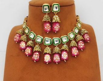 Peut inclure: Un ensemble collier et boucles d'oreilles doré. Le collier présente des accents carrés verts et blancs, avec des pendentifs roses en forme de cloche. Les boucles d'oreilles reprennent le design du collier. Les bijoux sont exposés sur un buste marron.