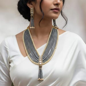May include: A multi-strand gray beaded necklace and matching earrings. The necklace features a gold pendant with clear stones and a tassel. The earrings have a similar design with a gold top and gray beaded tassels. The jewelry is worn over a white garment.