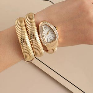 May include: A gold-tone quartz watch with a flexible, ribbed coil band that wraps around the wrist. The watch features a teardrop-shaped face with a white dial, gold hands, and a bezel accented with small clear stones around the perimeter.