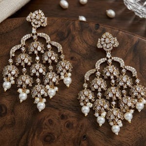 May include: A pair of gold-tone chandelier earrings featuring multiple bell-shaped dangles encrusted with clear crystals. Each earring is adorned with small white pearls at the base and a floral-shaped stud at the top, creating a layered design.