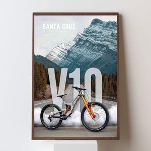 May include: Framed poster showing a Santa Cruz V10 mountain bike on a road with a mountain backdrop. The bike has orange and black details. The poster includes the text "SANTA CRUZ" and "V10".
