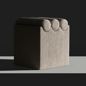 May include: A square, light brown upholstered ottoman with a textured surface. The top edge features three rounded, semi-circular details. The ottoman is set against a dark background with a white shelf.
