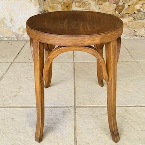 Op de afbeelding: Een ronde houten kruk met een medium bruine afwerking, voorzien van een cirkelvormige zitting en vier gebogen poten die met boogvormige steunen aan de onderkant van de zitting zijn bevestigd. De kruk heeft een glad, gepolijst oppervlak.