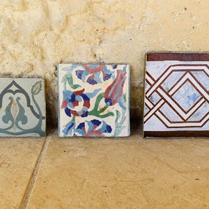 Può includere: Tre piastrelle quadrate in ceramica con motivi decorativi. Le piastrelle presentano disegni floreali e geometrici nei toni del blu, rosso, verde e marrone. Le piastrelle sono esposte su una superficie marrone chiaro.