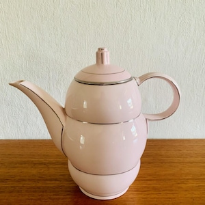 May include: A pale pink teapot with a rounded body, spout, and handle. The teapot has silver trim around the lid, body, and base. The lid has a small, cylindrical knob. The teapot is on a wooden surface.