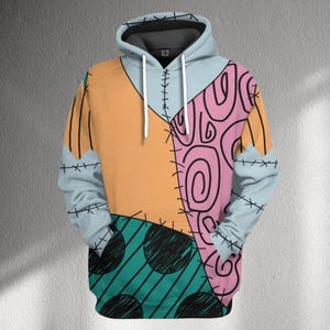 Könnte beinhalten: Ein Kapuzenpullover mit Patchwork-Design in Orange, Rosa, Türkis und Hellblau. Das Design umfasst schwarze Nähte und schwarze kreisförmige Muster. Die Ärmel und die Kapuze sind hellblau, im Stil eines Cartoon-Charakters.