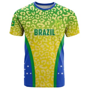 May include: A short-sleeved t-shirt with a vibrant design featuring the word "BRAZIL" in green block letters. The shirt has a yellow and green leopard print pattern on the top, with blue and white accents on the sides. A small Brazilian flag is visible.
