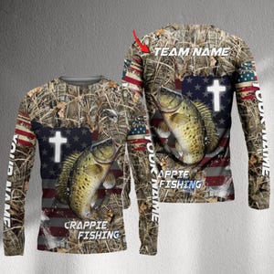 Op de afbeelding: Shirt met lange mouwen voor het vissen met een camouflagepatroon, een Amerikaans vlagontwerp en een crappie-visafbeelding. Het shirt heeft de tekst "CRAPPIE FISHING" en een wit kruis. De mouwen hebben de tekst "YOUR NAME" verticaal.