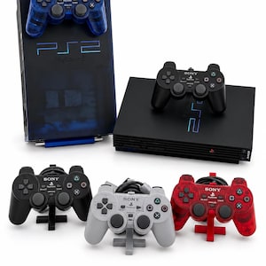 Op de afbeelding: Een verzameling PlayStation 2-consoles en controllers. De consoles zijn zwart, met de tekst "PS2" zichtbaar. Controllers zijn zwart, grijs, rood en blauw, met het Sony-logo. De controllers staan op standaards.