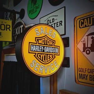 Op de afbeelding: Een rond, verlicht Harley-Davidson-bord met de woorden "SALES MOTOR HARLEY-DAVIDSON CYCLES SERVICE" in wit op een gele achtergrond. Andere borden zijn zichtbaar op de achtergrond.