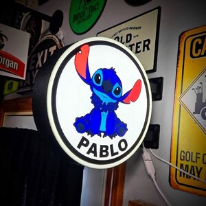 Op de afbeelding: Een ronde, verlichte bewegwijzering met het personage Stitch uit Lilo & Stitch. Het blauwe en rode personage staat op een witte achtergrond, met de naam "PABLO" in zwarte tekst eronder. De bewegwijzering heeft een zwarte rand.