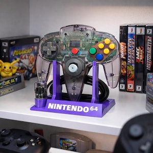 Könnte beinhalten: Ein transparenter Nintendo 64 Controller auf einem lila Ständer mit dem weißen Schriftzug "NINTENDO 64". Der Controller hat farbige Knöpfe und ein schwarzes Kabel. Weitere Videospiel-Zubehörteile sind im Hintergrund zu sehen.