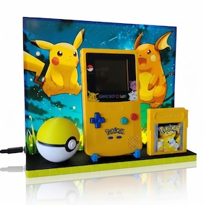 Könnte beinhalten: Gelbe Game Boy Color Konsole mit passender Spielkassette und Pokéball. Die Konsole zeigt Pikachu- und Raichu-Grafiken mit dem Text "GAME BOY COLOR" und "Pokémon". Der Hintergrund ist eine sternenklare Nachtszene.