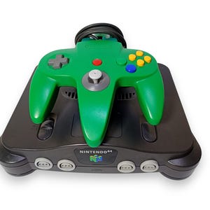 Può includere: Una console di gioco Nintendo 64 nera con un controller verde. Il controller ha un D-pad, un joystick analogico grigio e pulsanti colorati. La console presenta il logo Nintendo 64.