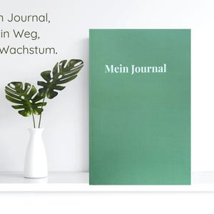 May include: A green journal with the text "Mein Journal" in white. The journal is next to a white vase with green leaves. The text "Dein Journal, dein Weg, dein Wachstum" is in the upper left corner.
