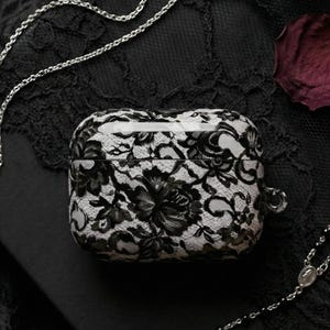 May include: A black and white floral lace AirPods Pro case sits on a black surface, with silver chains and a dark red rose petal. The case has a glossy finish and a small loop for attaching a keychain.