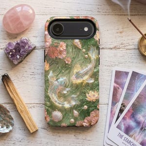 Green koi fish phone case with pink flowers on a white wood table beside rose quartz, amethyst, a wood incense stick and tarot cards.