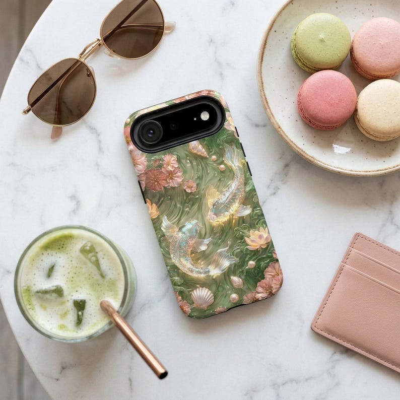 Green floral koi fish phone case on a white table beside sunglasses, iced matcha, pastel macarons and a small pink wallet.